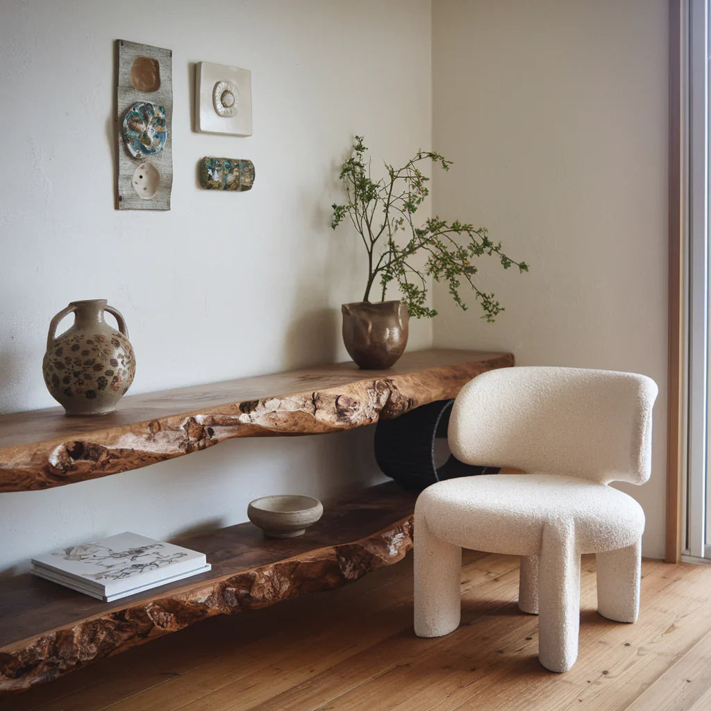 A photo of a Japandi-style room with a rustic wooden shelf and a minimalistic, white chair. There's a decorative ceramic pot and a plant on the shelf. The wall has a few pieces of artwork. The room has a wooden floor. 