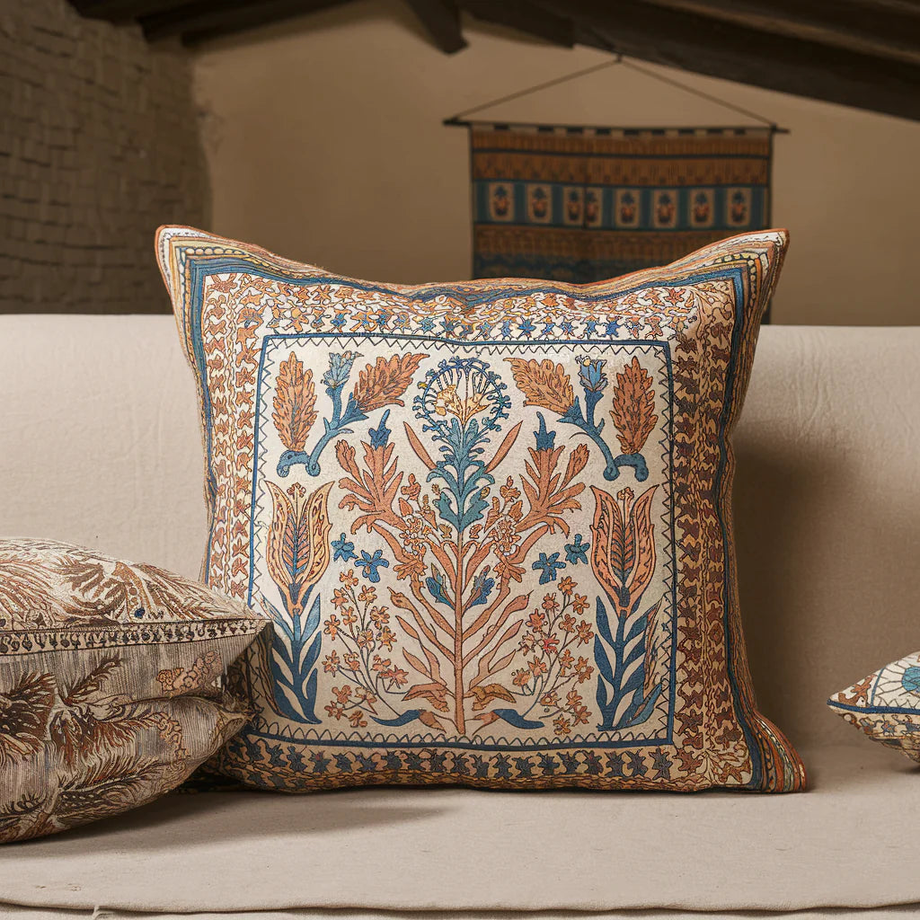 A photo of a hand block print décor item. The item is a large, square cushion with intricate hand-blocked patterns in orange, blue, and white. The background is a beige room with a few other hand block print décor items