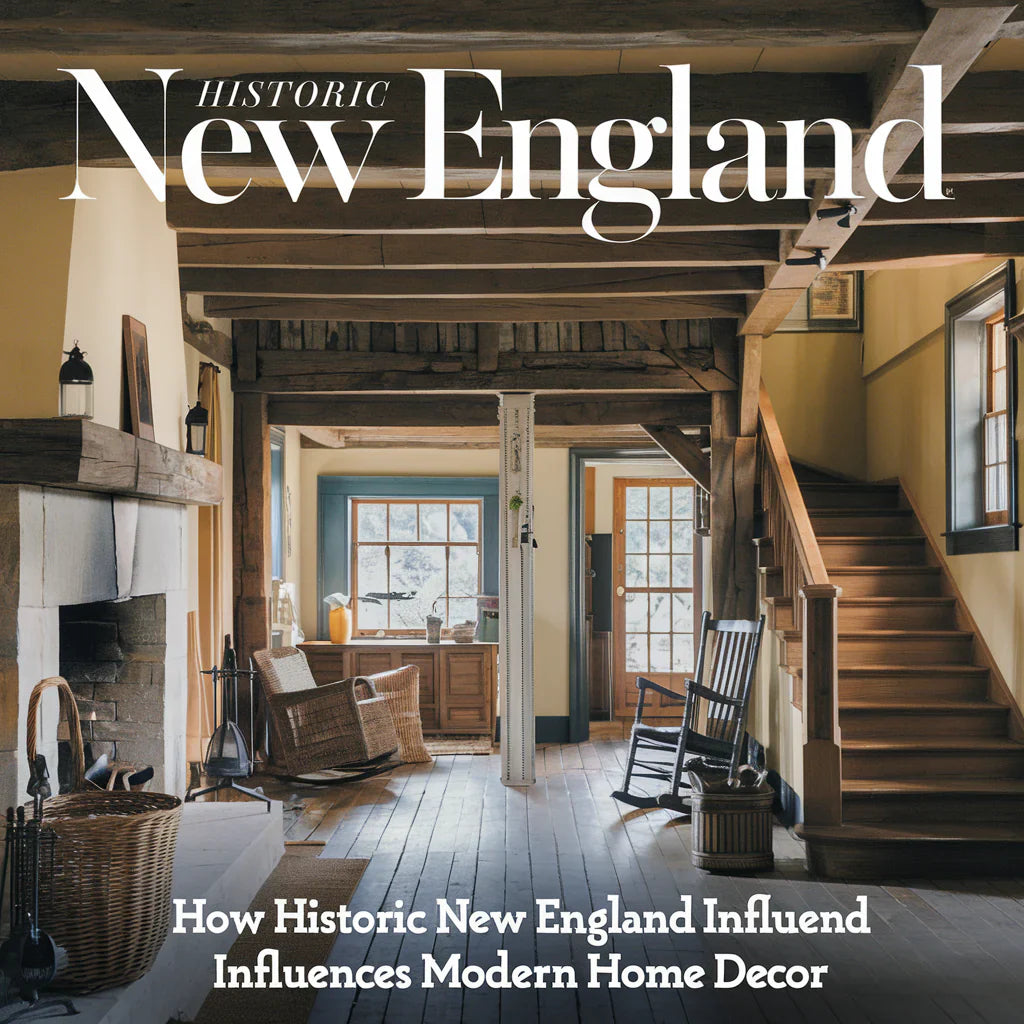 A photo of a historic New England home with rustic wood beams, a stone fireplace, and a wooden staircase. There are some vintage items like a lantern, a wicker basket, and a rocking chair.