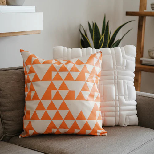 A photo of a home decor setting with two pillows on a couch. One pillow is made of latex and is bright orange. The other pillow is made of foam and is white. The latex pillow has a pattern of orange and white triangles