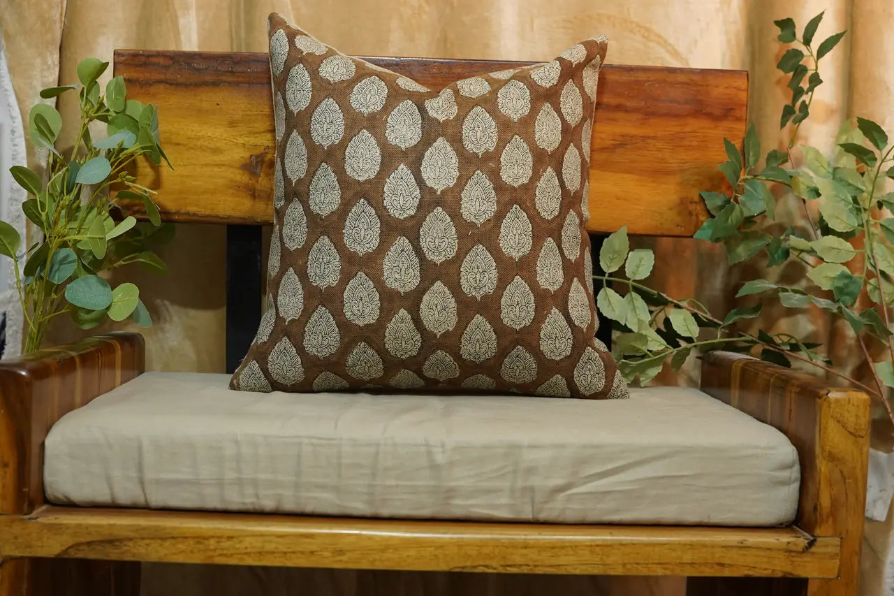 An indoor setting, possibly a living room or a lounge area. Dominating the scene is a wooden bench with a beige cushion. Atop the cushion is a decorative pillow with a repeating leaf pattern in brown and white.